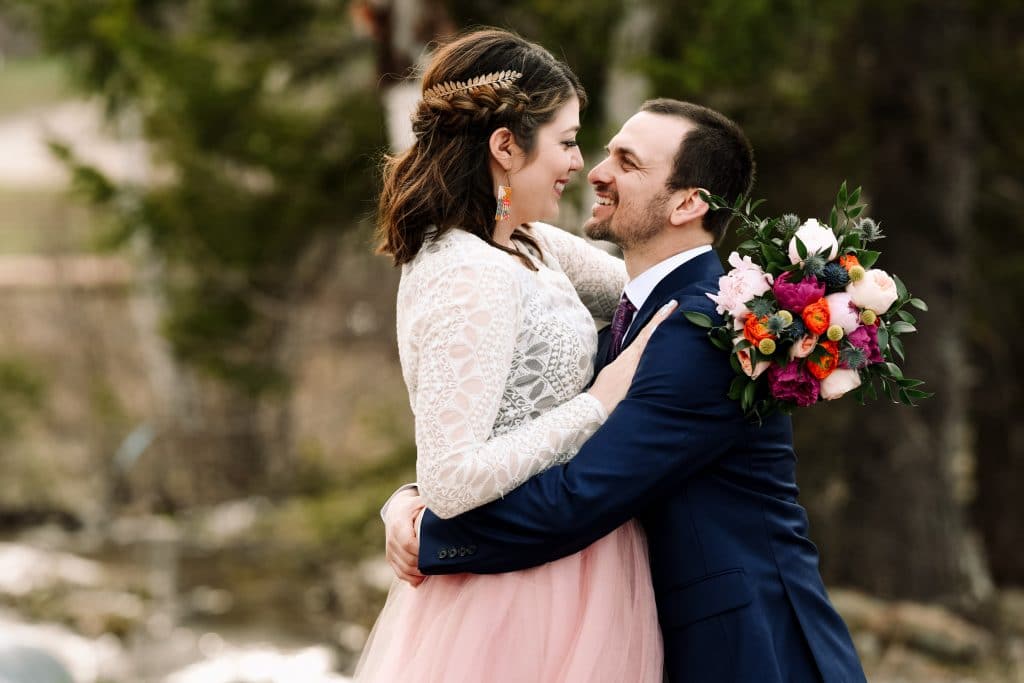 The bride and groom gaze into each others eyes as they embrace before a kiss. The bride has a leaf hair clasp and a colourful bouquet with sea holly flowers and pink peonies.