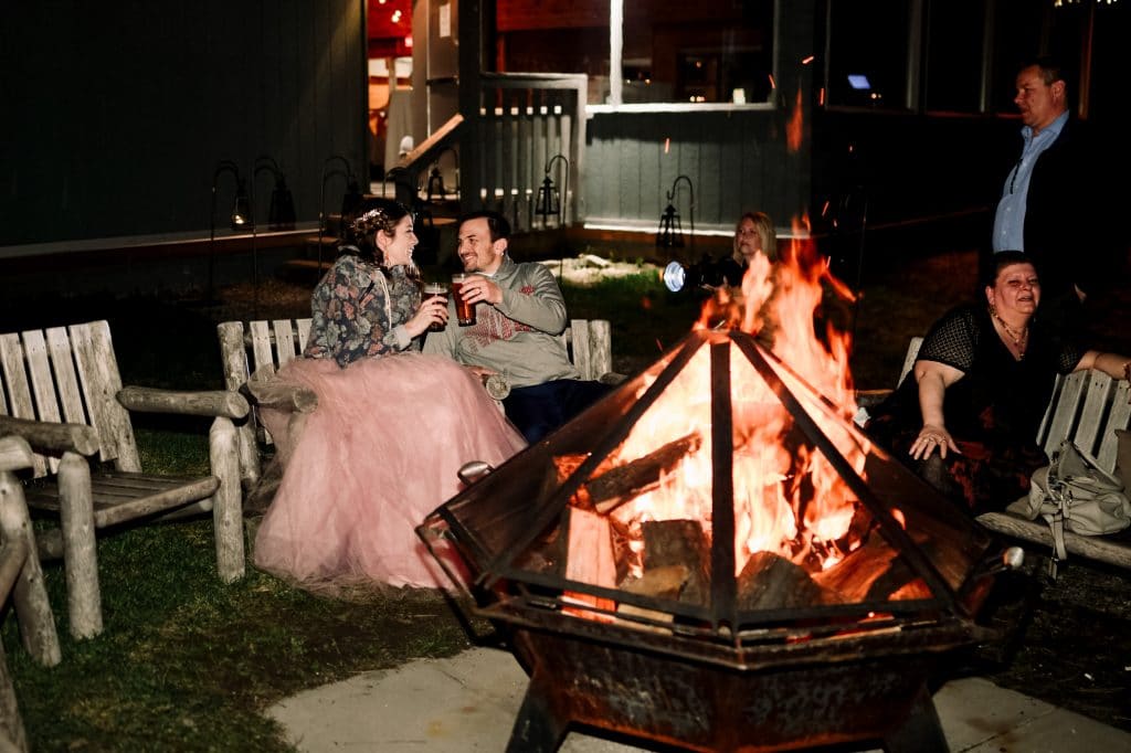 The bride and groom clink beer glasses while sitting outside at a camp fire on their wedding night.