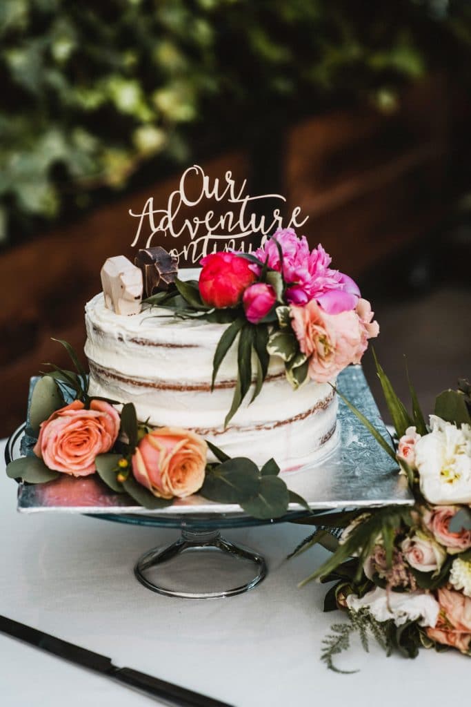 Small one-tier white wedding cake with fresh flowers and "our adventure continues" cake topper
