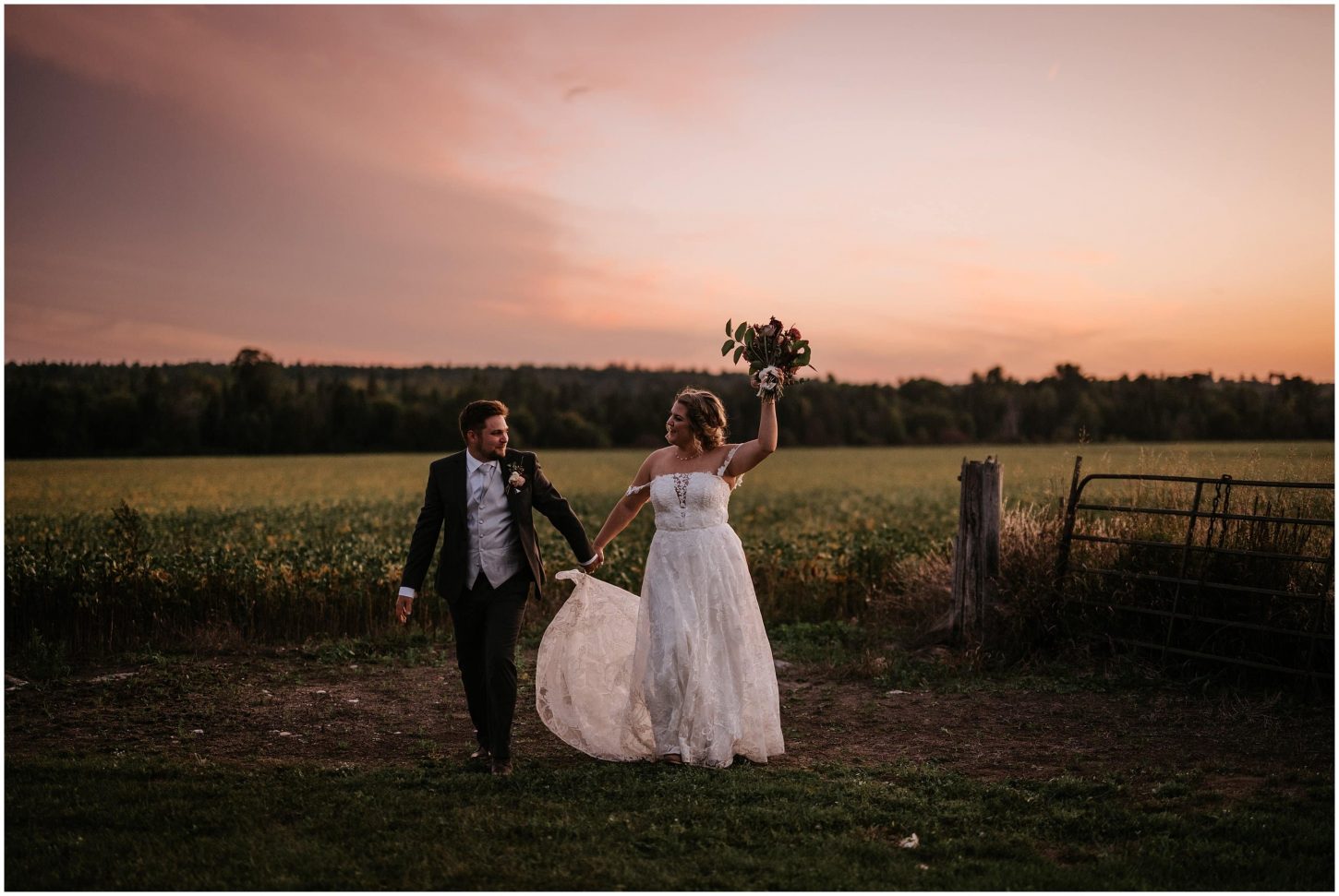 A bride and groom cheer in a field as the pink sun sets