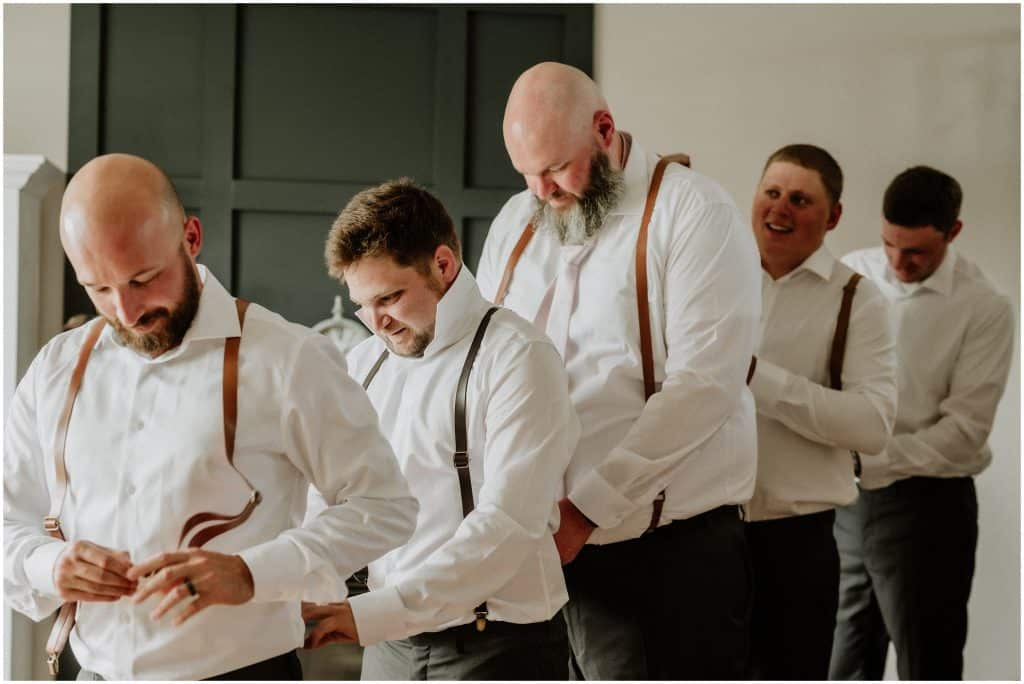 Groom and his groomsmen helping each other put on suspenders. Photo by Cindy Lottes Photography