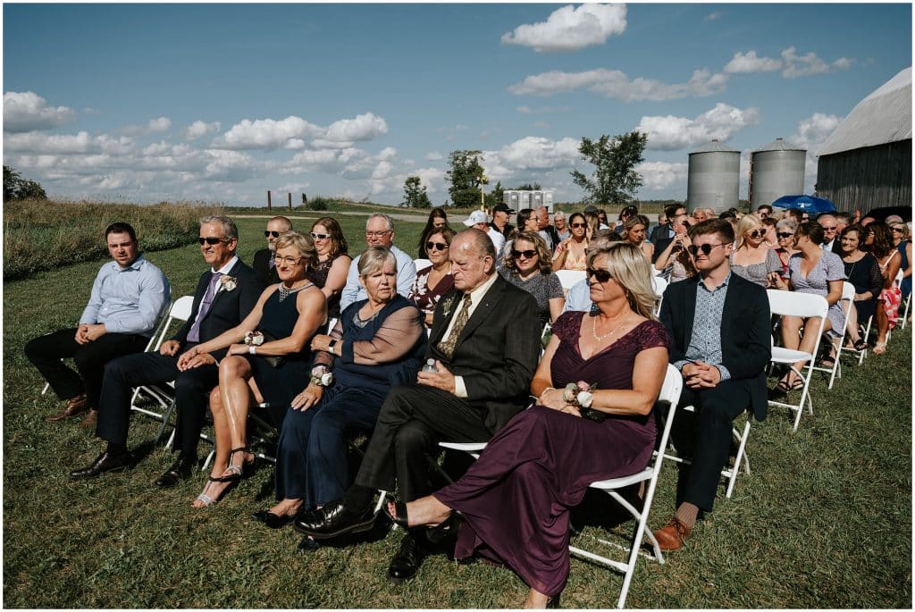 Friends and family sit on white chairs in a field during this outdoor September wedding. Photo by Cindy Lottes Photography
