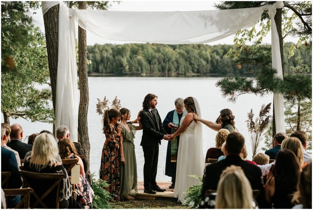 The bride and groom stand in front of a lake during their wedding ceremony and their friends and family help bless them.
